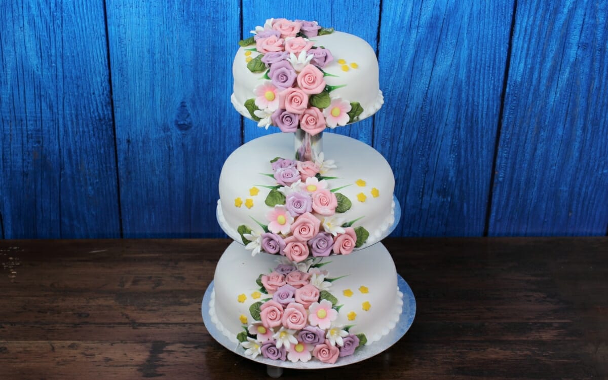 Bryllupskake i tre etasjer med pynt av roser og blomster i rosa, lilla og gult på hvitt marsipanlokk
