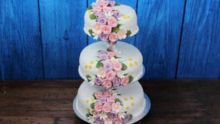 Bryllupskake i tre etasjer med pynt av roser og blomster i rosa, lilla og gult på hvitt marsipanlokk